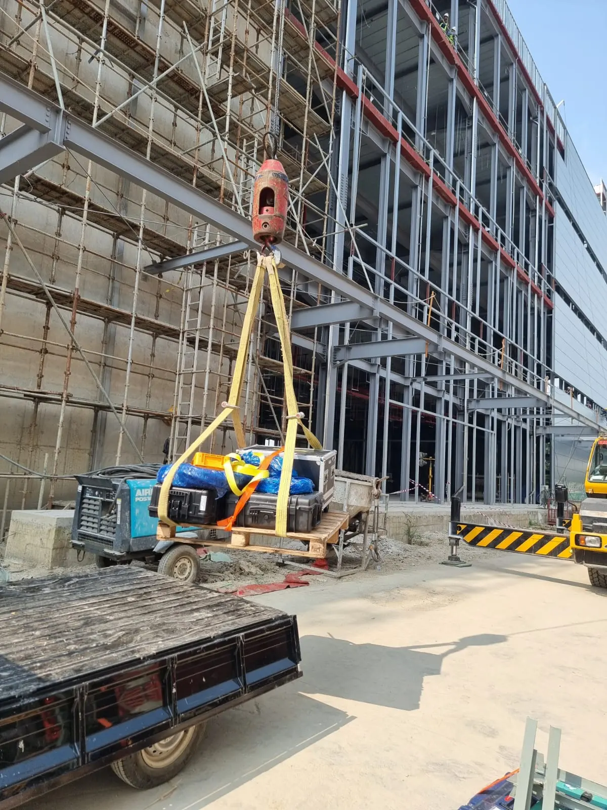Equipment cases rigged with yellow lifting slings and hoisted past a steel-frame factory building